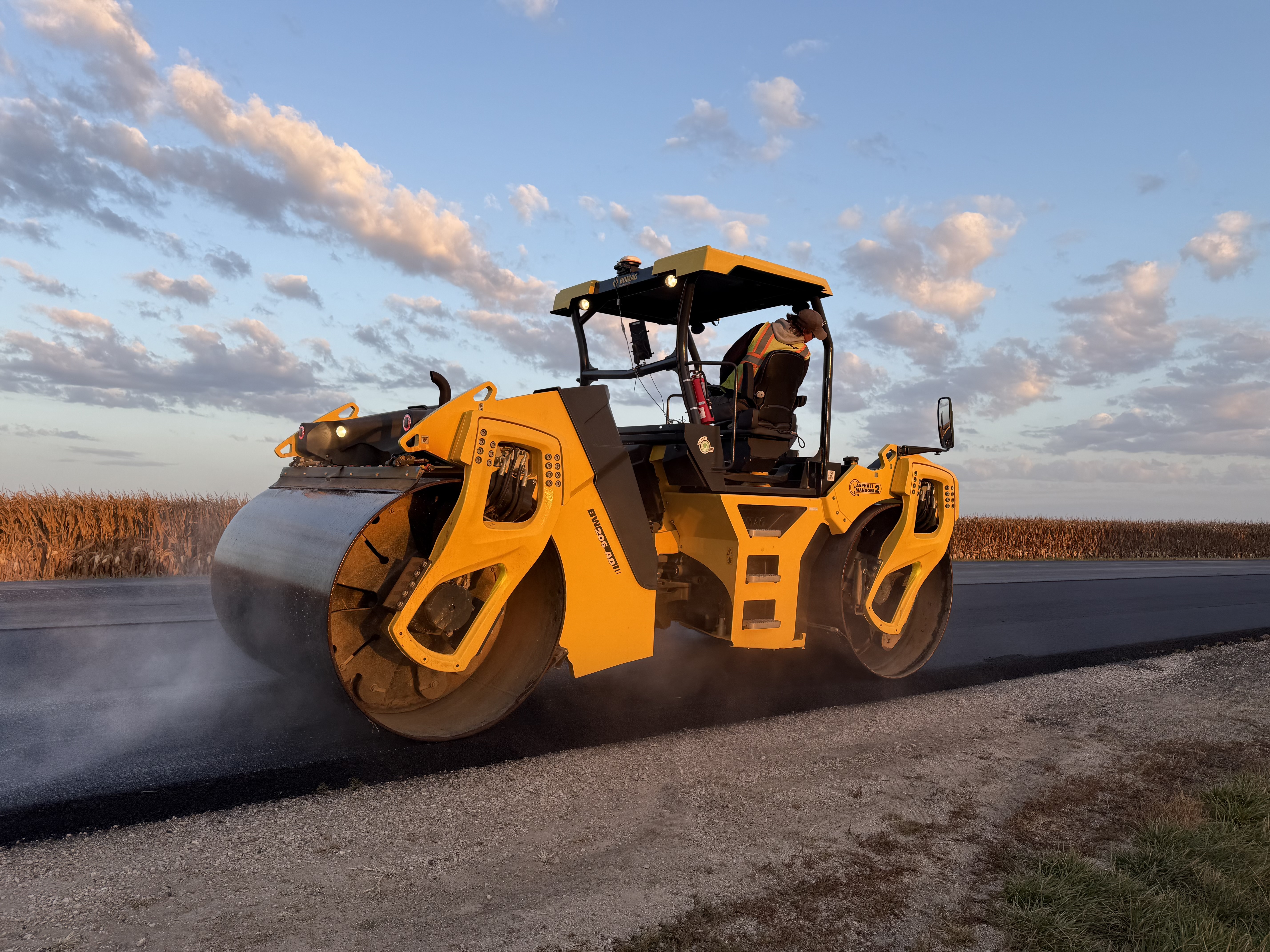 Yellow tandem asphalt roller compacting fresh pavement on a rural road with cornfields and cloudy sky in background