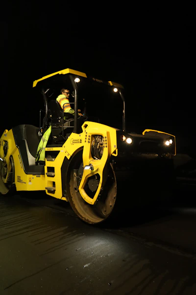 Yellow asphalt roller compactor operating at night with operator in cab and headlights illuminated