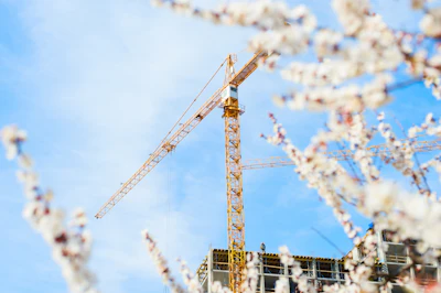 Yellow tower crane on construction site with white flowering branches in foreground and blue sky