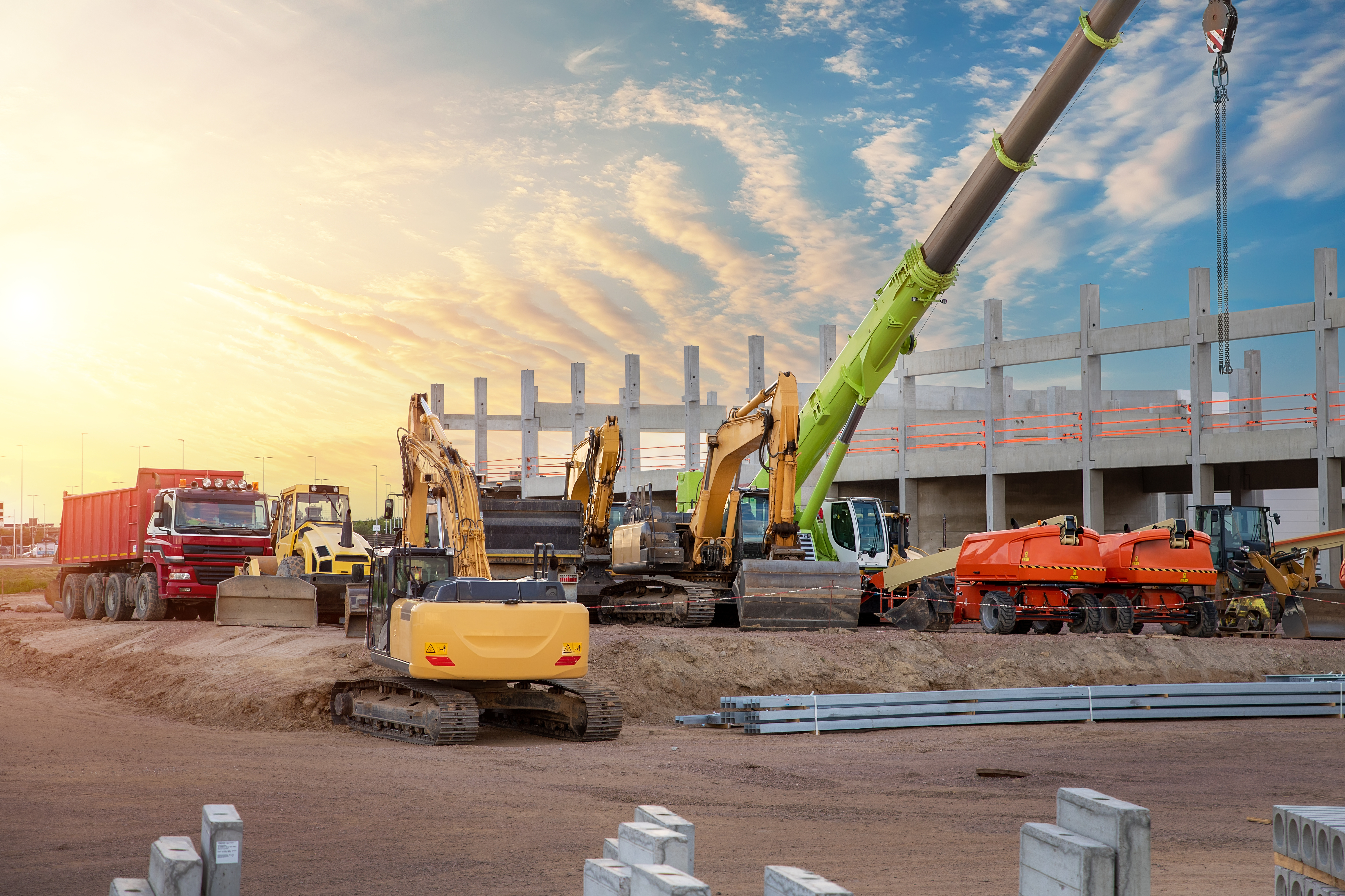 Construction site with heavy machinery, excavators, concrete pump, and trucks at sunset with building framework in background.