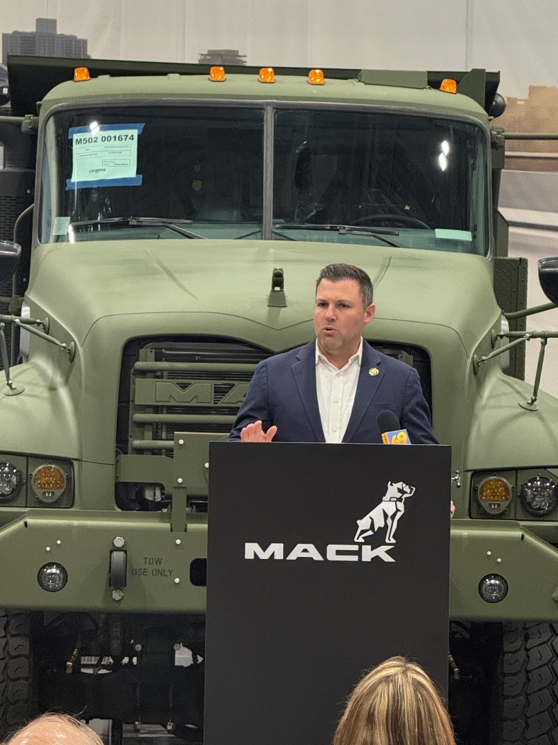 Man in business attire speaking at Mack Trucks podium in front of military truck