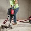 Construction worker operating a red and black Milwaukee jackhammer on concrete floor with compressor equipment nearby