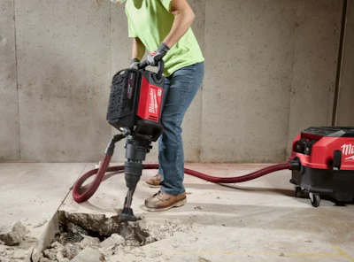 Construction worker operating a red and black Milwaukee jackhammer on concrete floor with compressor equipment nearby