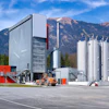 Industrial manufacturing facility with red building, silver storage tanks, and orange equipment against mountain backdrop