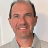 Smiling man with receding dark hair wearing a striped gray and white collared shirt against a white interior background