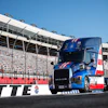 Blue and red Mack truck on racing track with stadium grandstands in background