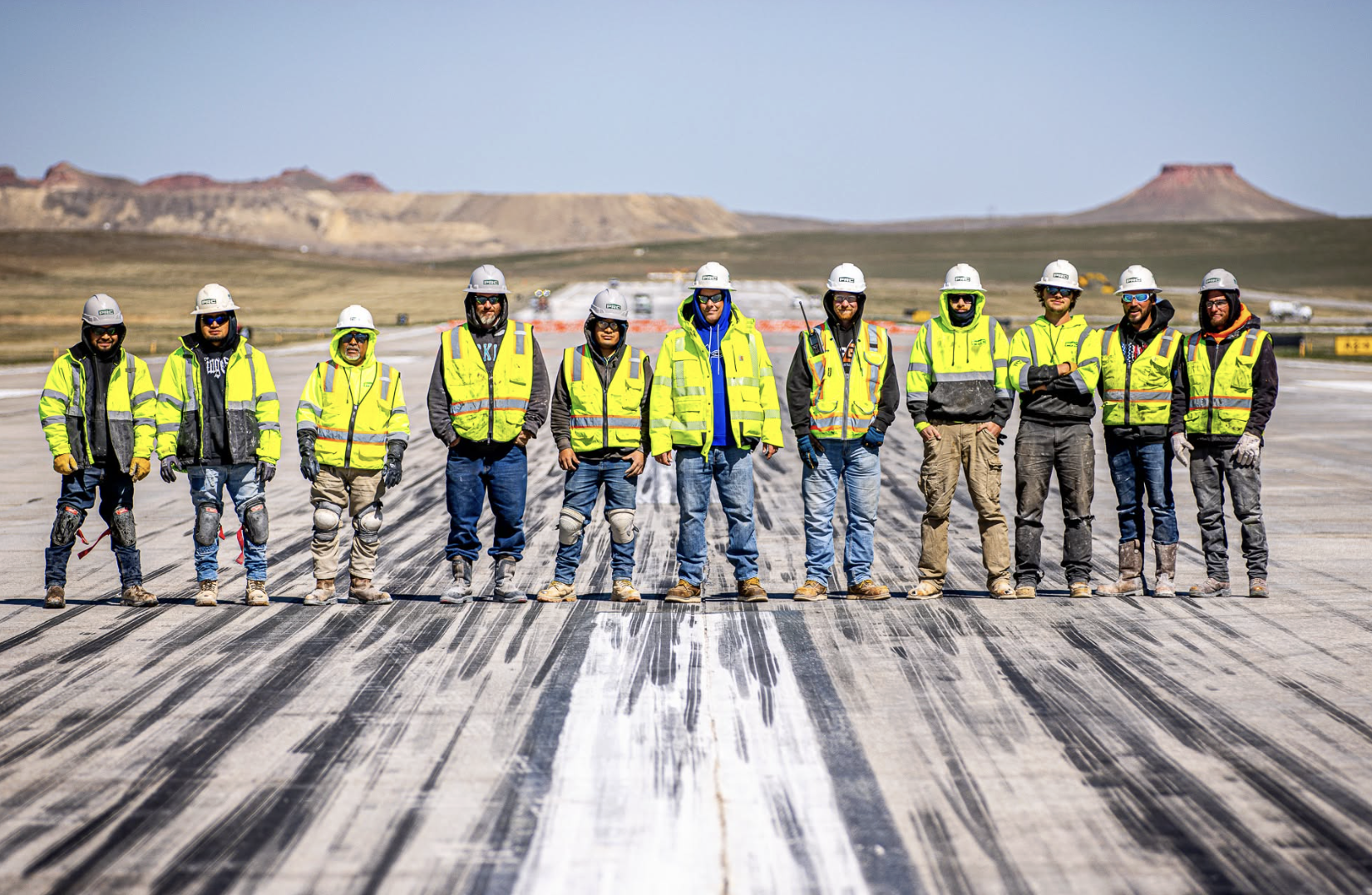 Team of construction workers in yellow safety vests and hard hats standing on airport runway with desert landscape
