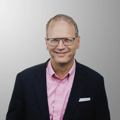 Professional headshot of a man wearing glasses, pink shirt, and dark blazer against neutral background