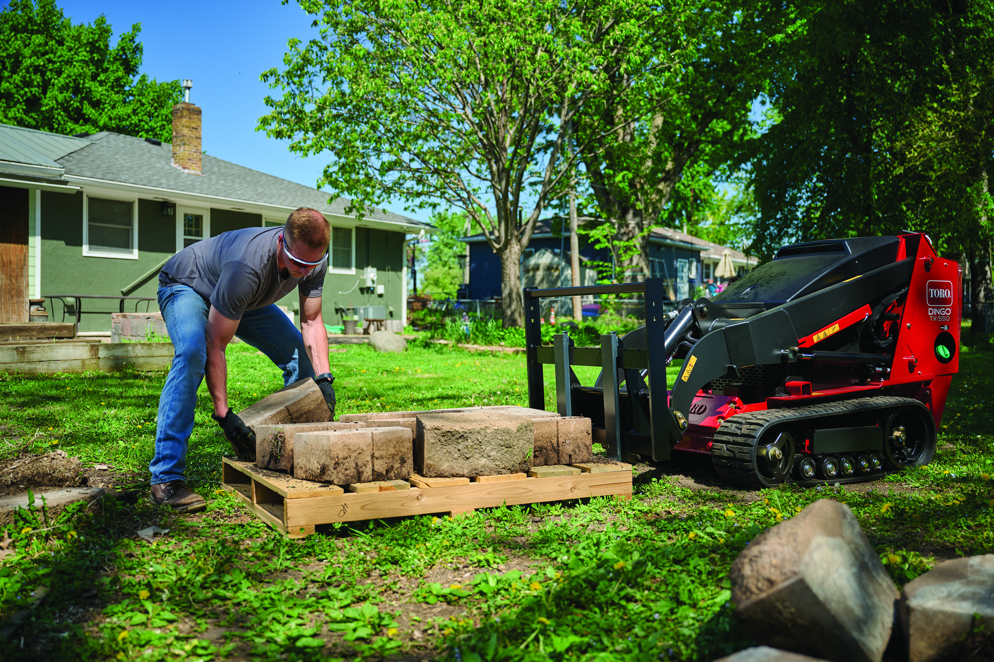 Man loading sod rolls onto a red Toro compact track loader in a residential yard with trees and houses.