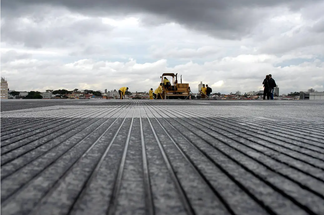 Construction crew operating heavy machinery on a large flat surface with parallel ridged patterns under cloudy sky.