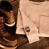 Brown leather work boots with cream crew neck sweater and watch arranged on wooden surface