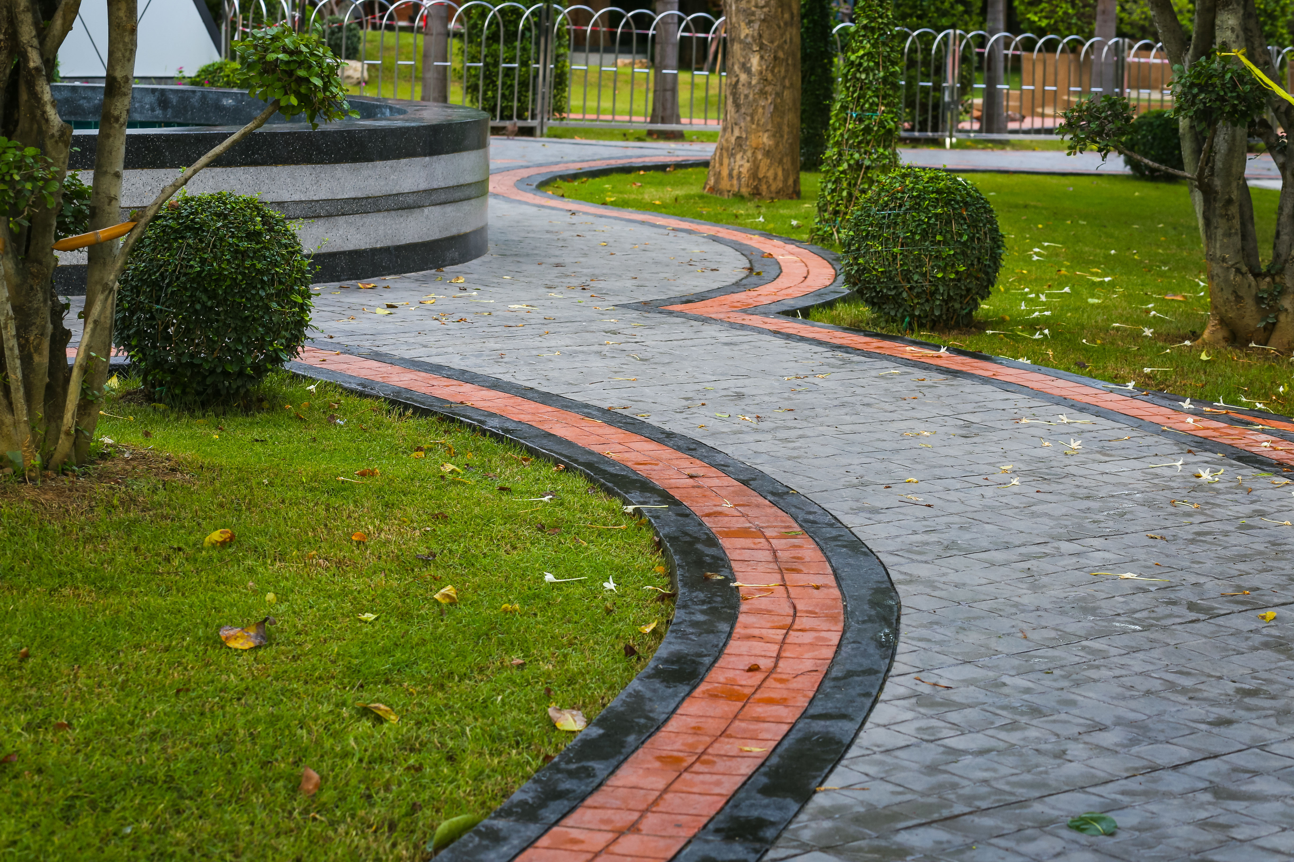 Curved brick and stone pathway winding through manicured park with green lawn, topiary bushes, and mature trees.