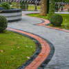 Curved brick and stone pathway winding through manicured park with green lawn, topiary bushes, and mature trees.