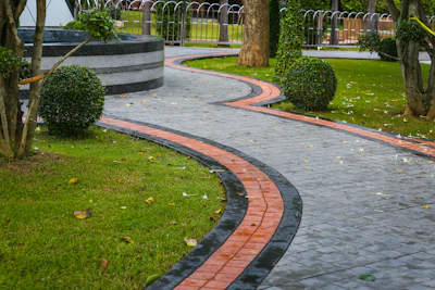 Curved brick and stone pathway winding through manicured park with green lawn, topiary bushes, and mature trees.