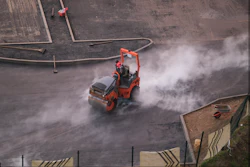 Red vibratory roller compactor creating dust clouds while compacting asphalt on construction site, aerial view