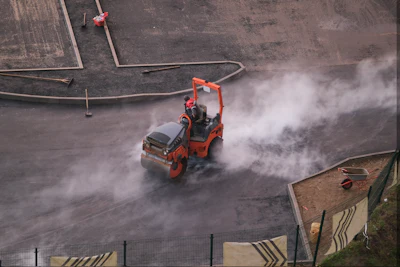 Red vibratory roller compactor creating dust clouds while compacting asphalt on construction site, aerial view