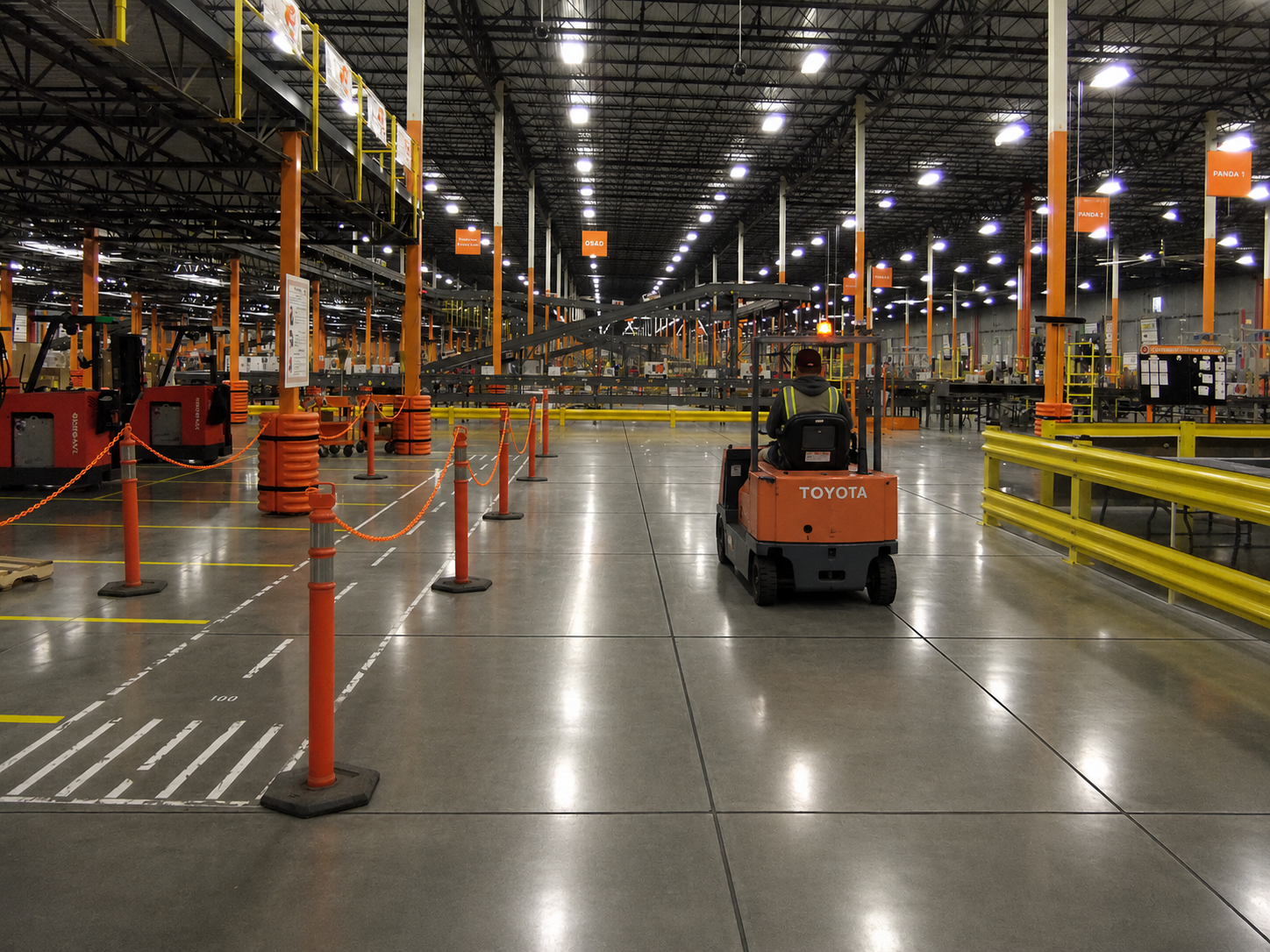 Industrial warehouse with Toyota forklift navigating between red safety barriers and yellow guardrails under bright LED lighting.