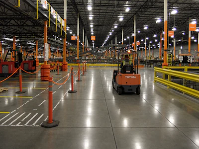 Industrial warehouse with Toyota forklift navigating between red safety barriers and yellow guardrails under bright LED lighting.