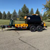 Yellow and black dual-axle asphalt tanker truck parked on pavement with evergreen trees and American flag in background