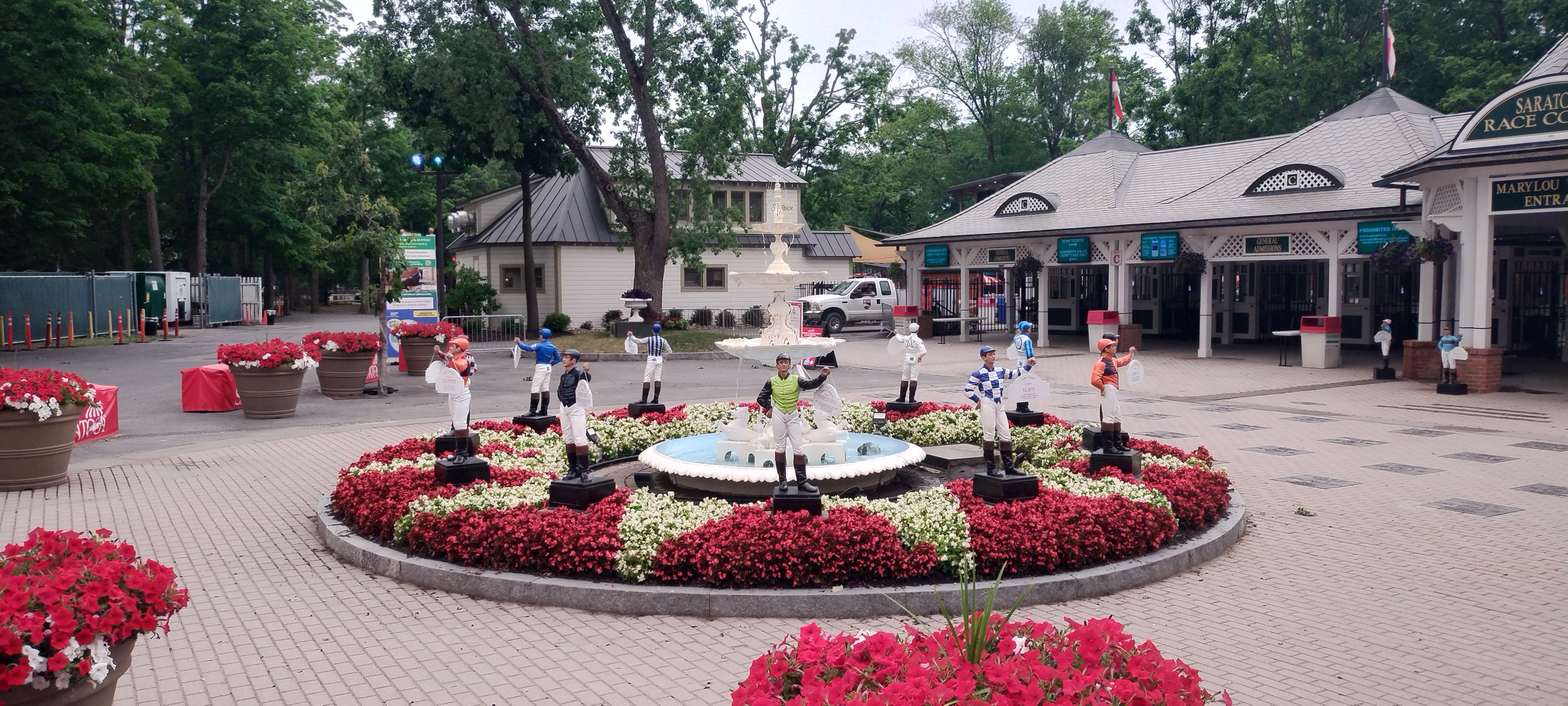 Vibrant plaza courtyard with circular red and white floral garden surrounding central fountain, bordered by shops and trees