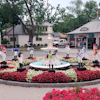 Vibrant plaza courtyard with circular red and white floral garden surrounding central fountain, bordered by shops and trees