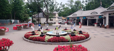 Vibrant plaza courtyard with circular red and white floral garden surrounding central fountain, bordered by shops and trees