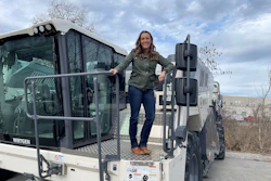 Woman posing on Wirtgen industrial truck with tire racks and bare landscape in background
