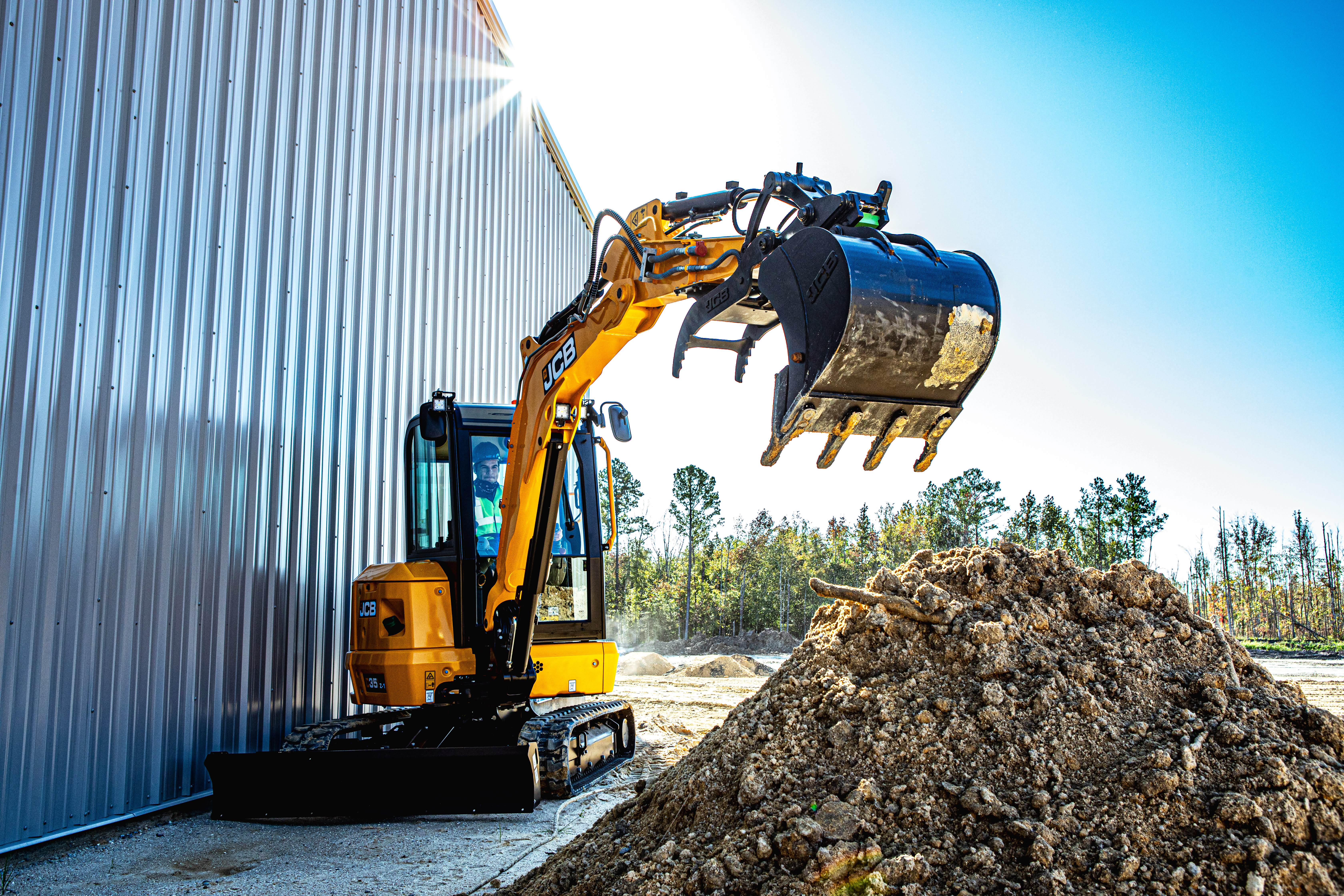 Yellow and black compact excavator loading soil into a blue bucket near a corrugated metal building on a sunny day