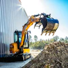 Yellow and black compact excavator loading soil into a blue bucket near a corrugated metal building on a sunny day