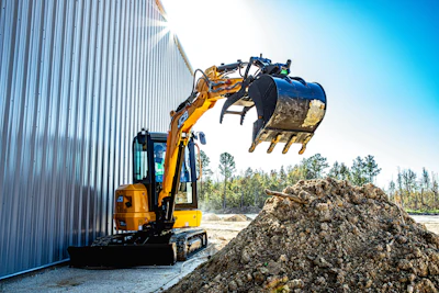 Yellow and black compact excavator loading soil into a blue bucket near a corrugated metal building on a sunny day