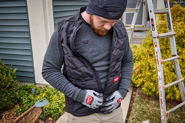 Worker wearing black Milwaukee branded vest and gloves standing near ladder and garden plants