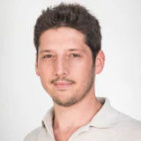Professional headshot of a man with dark hair and facial hair wearing a light-colored collared shirt against a neutral background