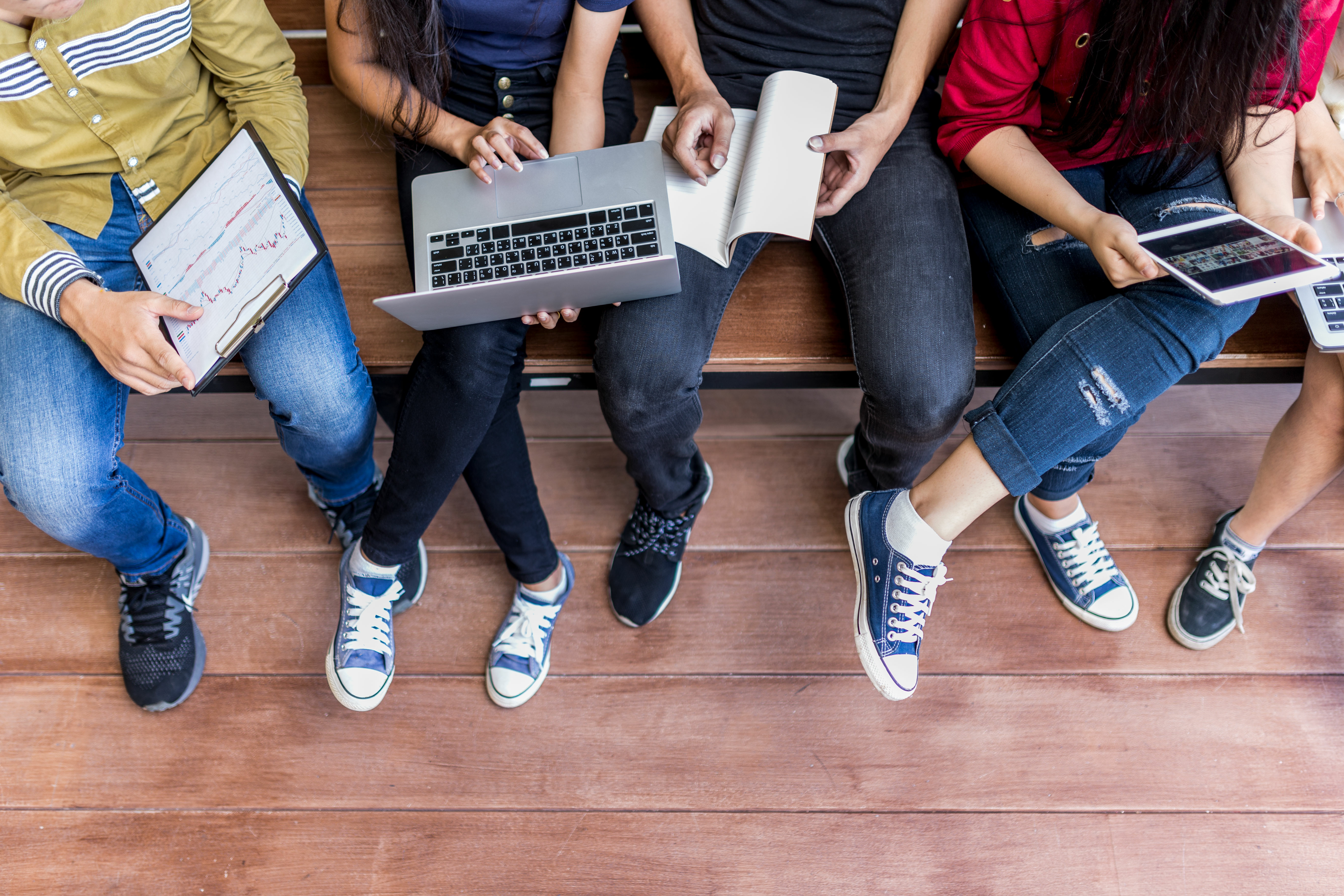 Group of young people sitting together on wooden floor collaborating with laptop and mobile devices