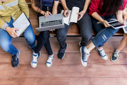 Group of young people sitting together on wooden floor collaborating with laptop and mobile devices