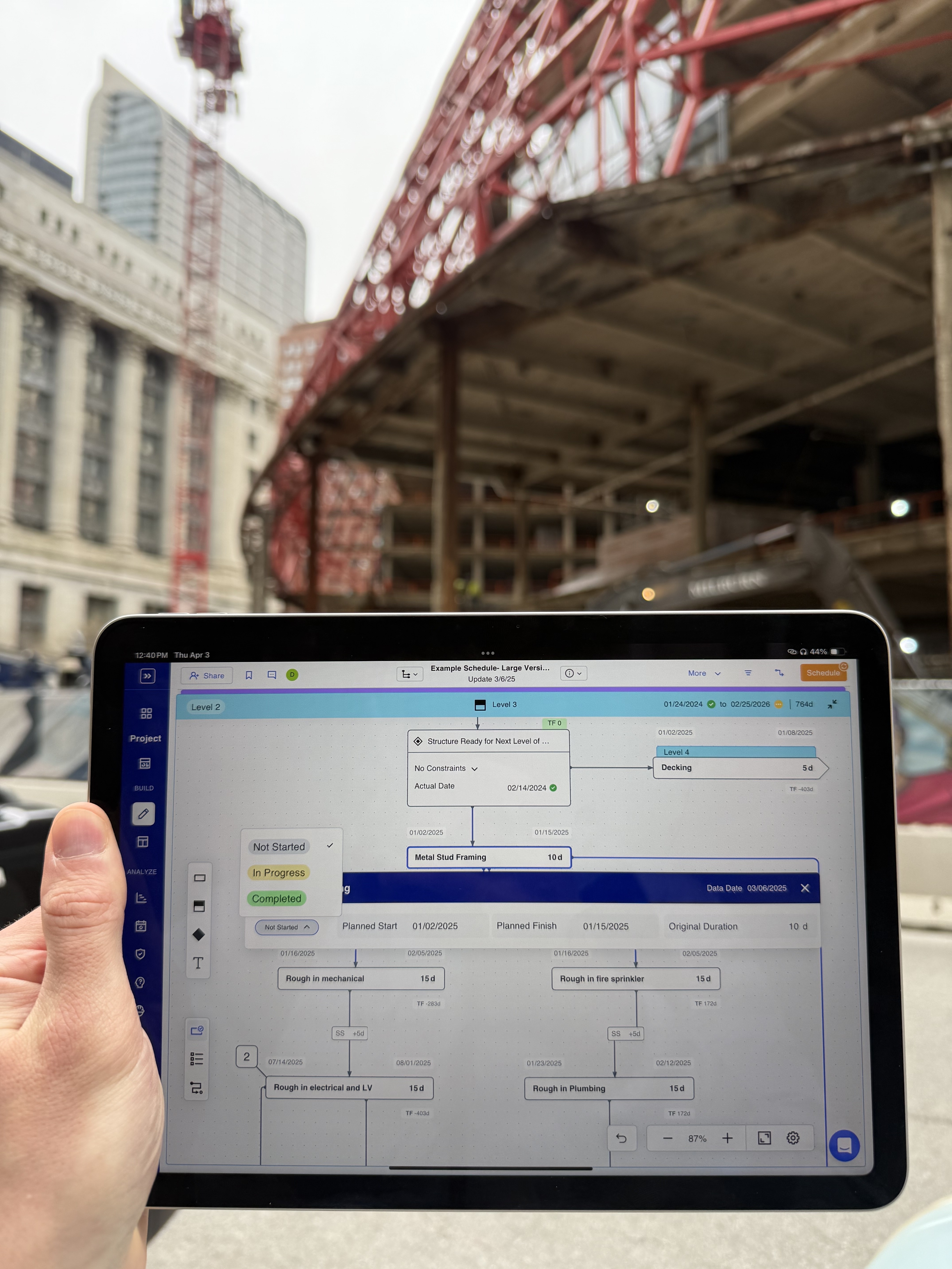 Person holding tablet displaying construction project schedule with Gantt chart, urban bridge and buildings in background