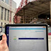 Person holding tablet displaying construction project schedule with Gantt chart, urban bridge and buildings in background