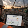 Construction worker reviewing project plans on tablet at railway bridge construction site during sunset