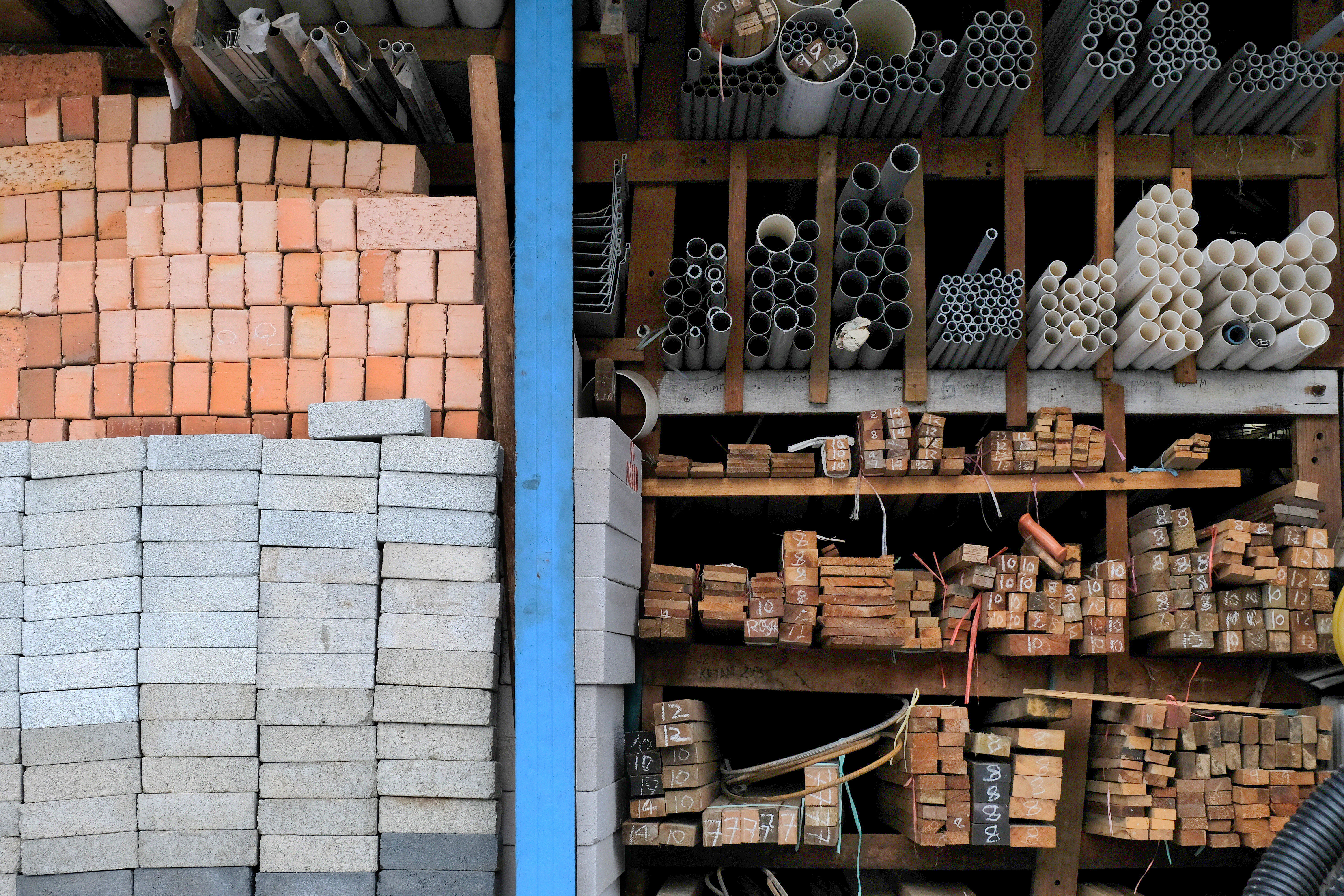 Construction materials warehouse with stacked red bricks, white concrete blocks, metal pipes, and wooden lumber organized on shelves.