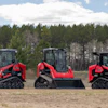 Three red and black tracked skid steer loaders lined up in a field with forest background
