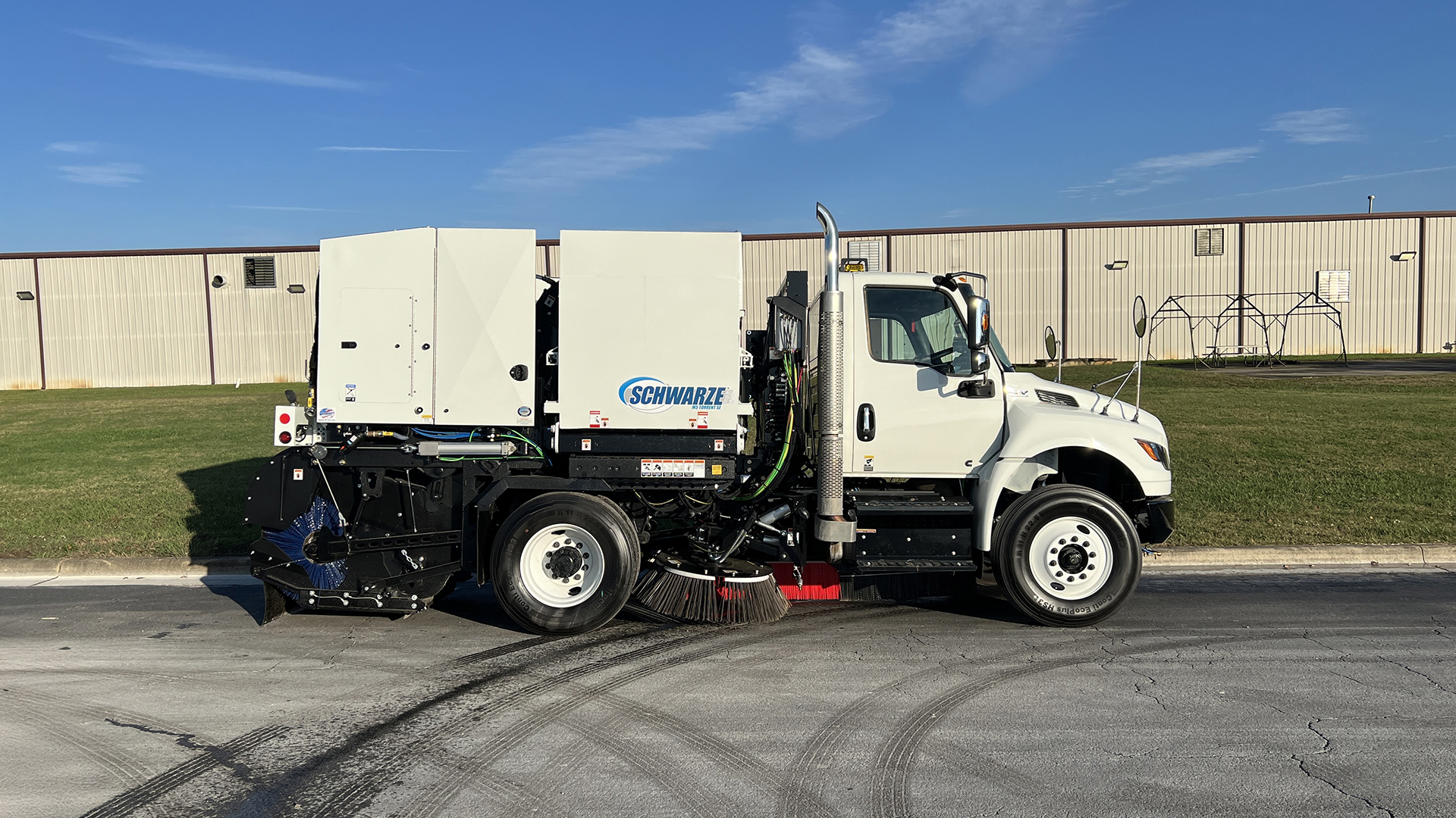 White Schwarze street sweeper truck with blue and black mechanical broom equipment parked on asphalt near industrial buildings