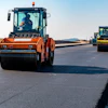 Road construction crew operating orange and yellow asphalt rollers on a newly paved highway under clear skies.