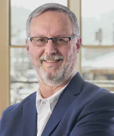 Professional headshot of a bearded man wearing glasses and navy blazer in modern office setting