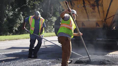 Road construction workers in safety vests operating asphalt paving equipment on a residential street with a paving truck nearby.