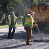 Road construction workers in safety vests operating asphalt paving equipment on a residential street with a paving truck nearby.
