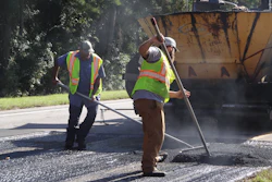 Road construction workers in safety vests operating asphalt paving equipment on a residential street with a paving truck nearby.
