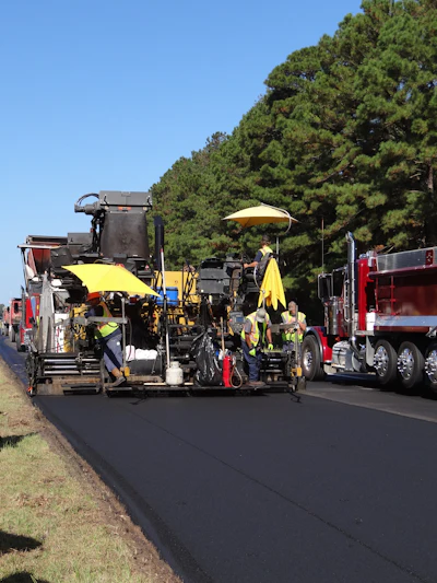 Road construction crew operating asphalt paving equipment with yellow umbrellas for sun protection on a sunny day