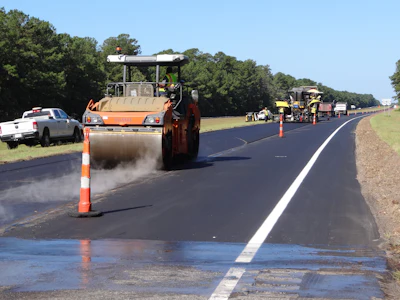 Asphalt paving roller compacting fresh pavement during highway construction with work crew and equipment