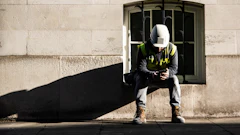 Construction worker in safety gear sitting against stone building wall in sunlight