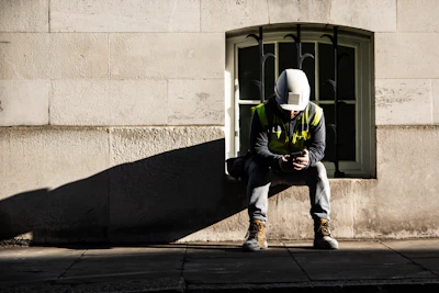 Construction worker in safety gear sitting against stone building wall in sunlight