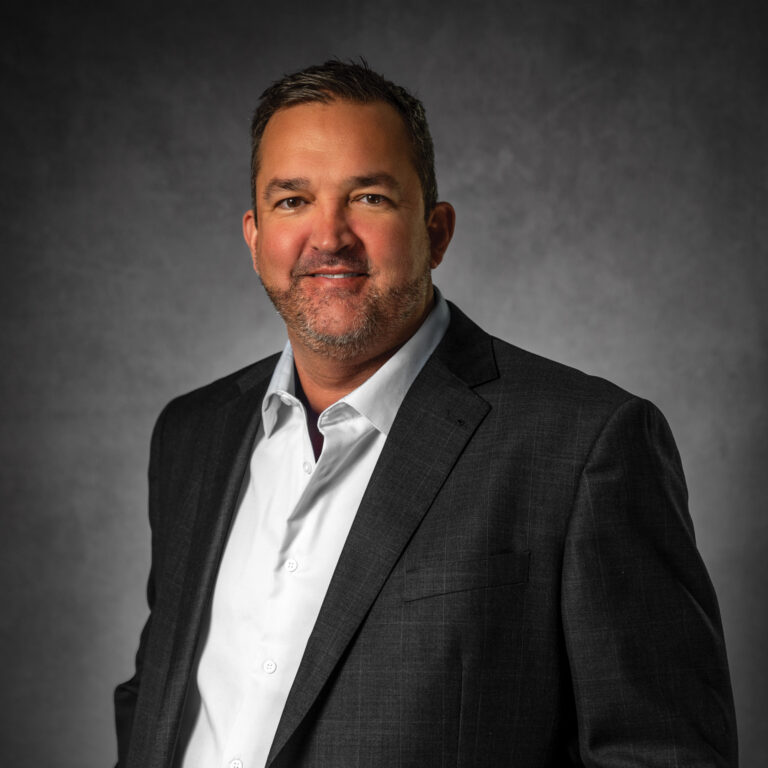 Professional headshot of a man in dark blazer and white shirt against gray background Headshot